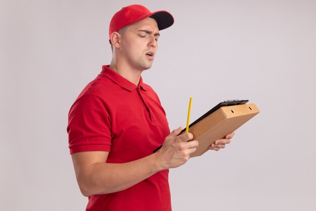 Confused young delivery man wearing uniform with cap holding clipboard with pizza box - isolated on white wall