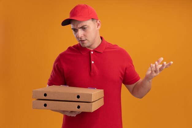 Confused young delivery man wearing uniform with cap holding and looking at pizza boxes spreading hand isolated on orange wall