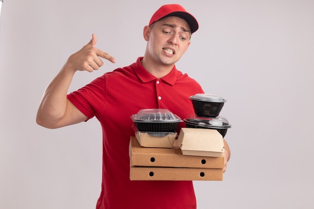 Смущенный молодой курьер в униформе с крышкой и указывает на контейнеры для еды на коробках для пиццы, изолированных на белой стене