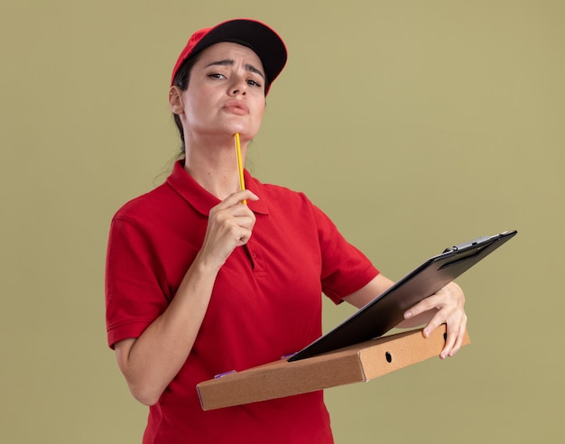 オリーブグリーンの壁に分離された鉛筆で顎に触れるピザパッケージクリップボードを保持している制服と帽子で混乱した若い配達の女性