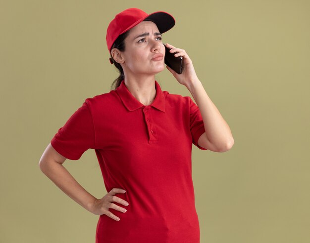 Confused young delivery woman in uniform and cap keeping hand on waist talking on phone looking at side 