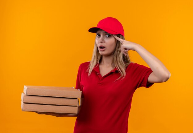 Confused young delivery woman wearing red uniform and cap holding pizza box putting finger on forehead 