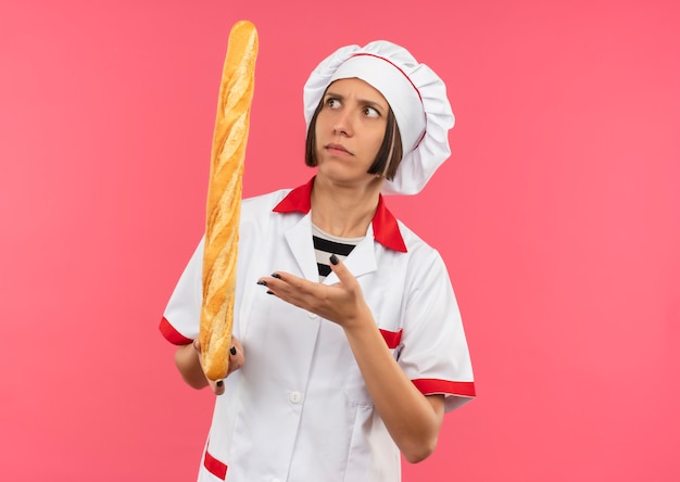 Смущенная молодая женщина-повар в униформе шеф-повара смотрит и показывает рукой на хлебную палочку, изолированную на розовом, с копией пространства