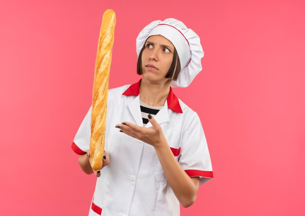 Confused young female cook in chef uniform holding looking and pointing with hand at bread stick isolated on pink  with copy space