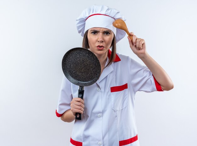 Confused young female cook wearing chef uniform holding frying pan with spoon isolated on white wall