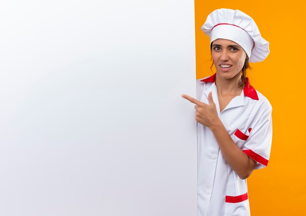 Confused young female cook wearing chef uniform holding and pointa to white wall on isolated yellow wall with copy space