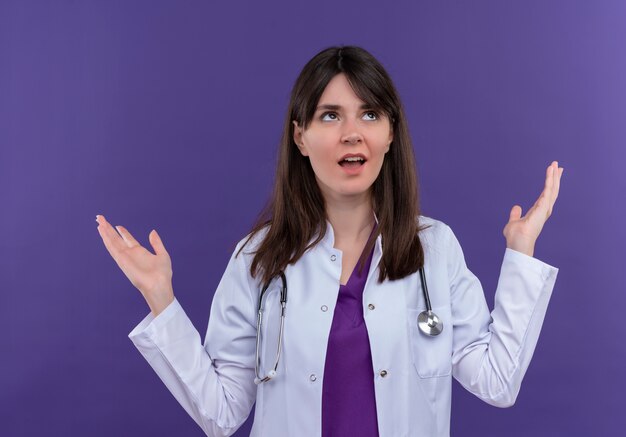Смущенная молодая женщина-врач в медицинском халате со стетоскопом держит руки вверх на изолированном фиолетовом фоне с копией пространства