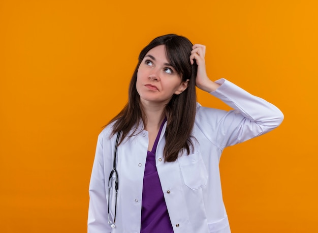 Confused young female doctor in medical robe with stethoscope puts hand on head on isolated orange background with copy space