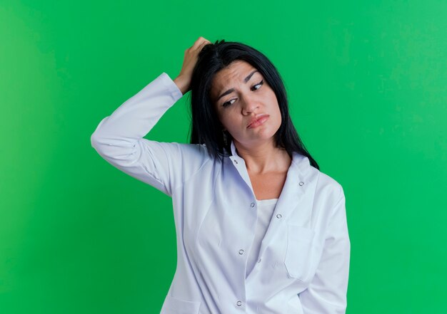 Confused young female doctor wearing medical robe looking at side touching head 