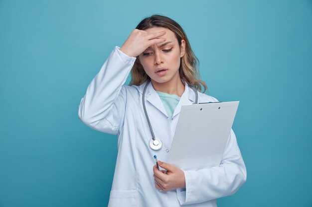 Confused young female doctor wearing medical robe and stethoscope around neck holding pen and clipboard keeping hand on head looking down 