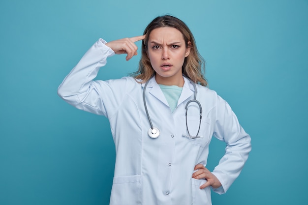 Confused young female doctor wearing medical robe and stethoscope around neck keeping hand on waist doing think gesture 