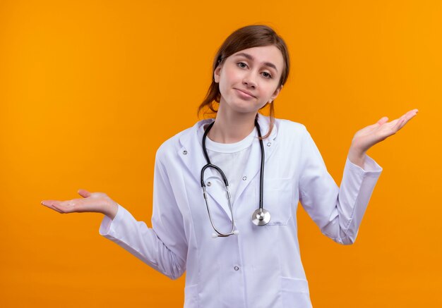 Confused young female doctor wearing medical robe and stethoscope showing empty hands on isolated orange space