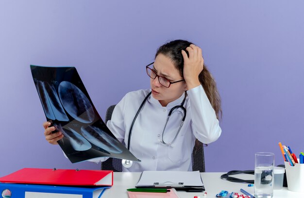 Confused young female doctor wearing medical robe and stethoscope sitting at desk with medical tools holding and looking at x-ray shot keeping hand on head isolated