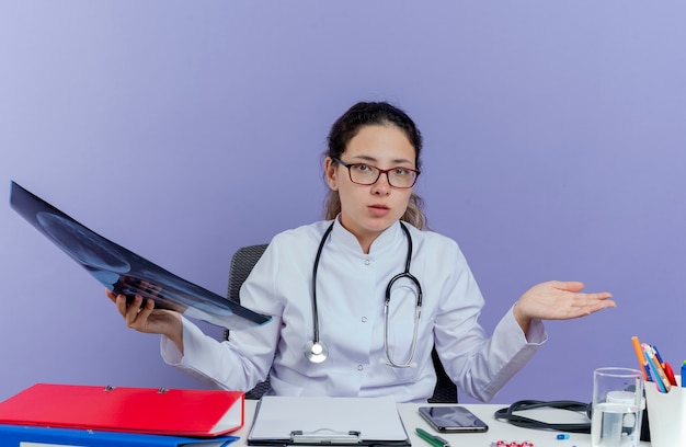 Free photo confused young female doctor wearing medical robe and stethoscope sitting at desk with medical tools holding x-ray shot looking showing empty hand isolated