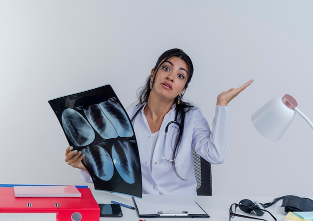 Смущенная молодая женщина-врач в медицинском халате и стетоскопе сидит за столом с медицинскими инструментами, держа рентгеновский снимок, глядя, показывая пустую изолированную руку