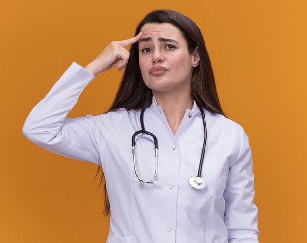 Confused young female doctor wearing medical robe with stethoscope puts finger on forehead looking at camera isolated on orange wall with copy space