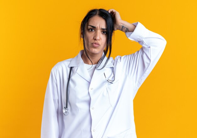 Confused young female doctor wearing medical robe with stethoscope scratching head isolated on yellow wall