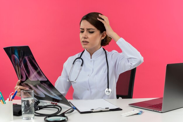 Смущенная молодая женщина-врач в медицинском халате со стетоскопом, сидя за столом, работает на компьютере, держа и глядя на рентгеновский снимок, положив руку на голову на розовой стене с копией пространства