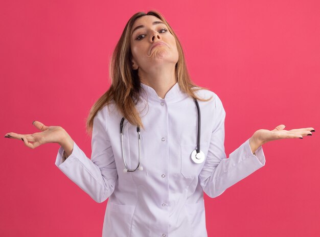 청진 기 핑크 벽에 고립 된 손을 확산 의료 가운을 입고 혼란 된 젊은 여성 의사