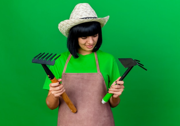 Confused young female gardener in uniform wearing gardening hat holds rake and hoe rake