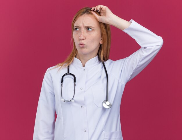 Confused young female ginger doctor wearing medical robe and stethoscope touching head looking at side with pursed lips isolated on crimson wall