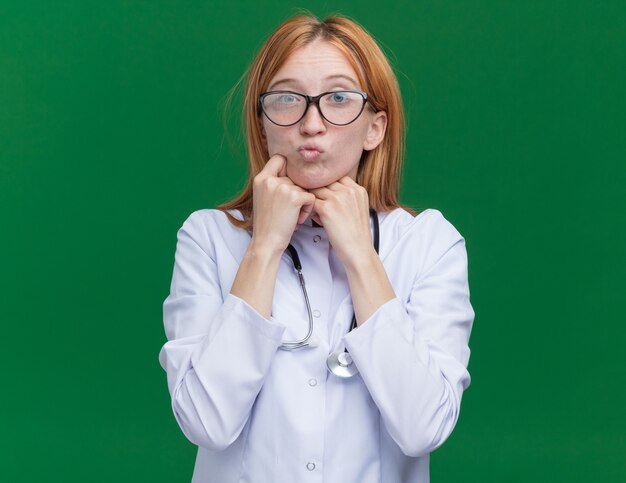 Confused young female ginger doctor wearing medical robe and stethoscope with glasses keeping hands under chin pursing lips 