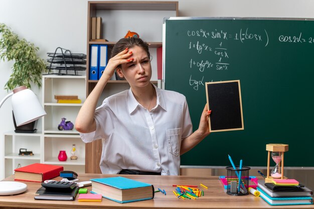 机に座って、教室の横を見て頭に手を置いてミニ黒板を持っている学用品と混乱している若い女性の数学の先生