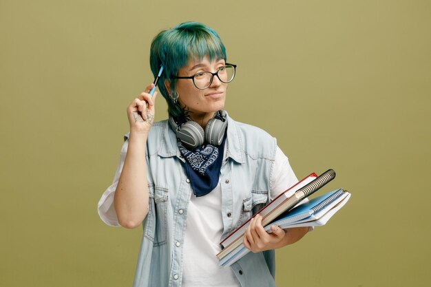 안경 반다나와 헤드폰을 목에 두른 혼란스러운 젊은 여학생은 올리브 녹색 배경에 격리된 펜으로 머리를 만지는 쪽을 바라보는 메모장을 들고 있습니다.