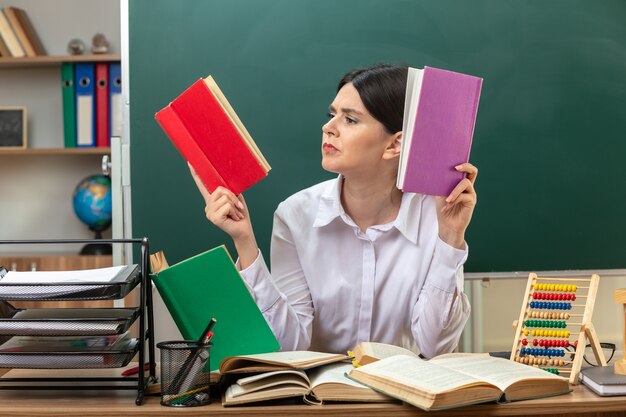 Confused young female teacher holding and reading book sitting at table with school tools in classroom