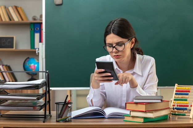 眼鏡をかけて、教室で学校の道具を持ってテーブルに座っている電卓を見て混乱している若い女性教師