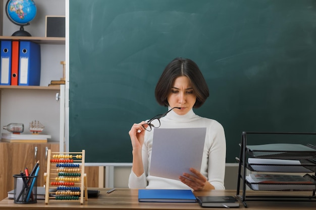 смущенная молодая учительница в очках, держащая бумагу, сидящая за партой со школьными инструментами в классе