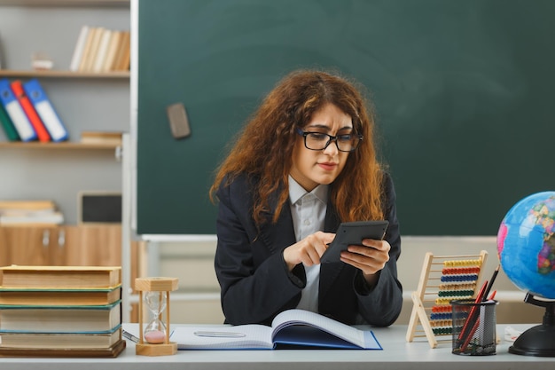 confused young female teacher wearing glasses holding and points at calculator sitting at desk with school tools in classroom