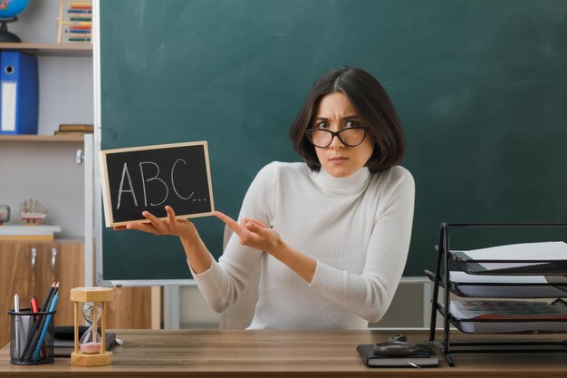 confused young female teacher wearing glasses holding and points at mini chalkboard sitting at desk with school tools on in classroom