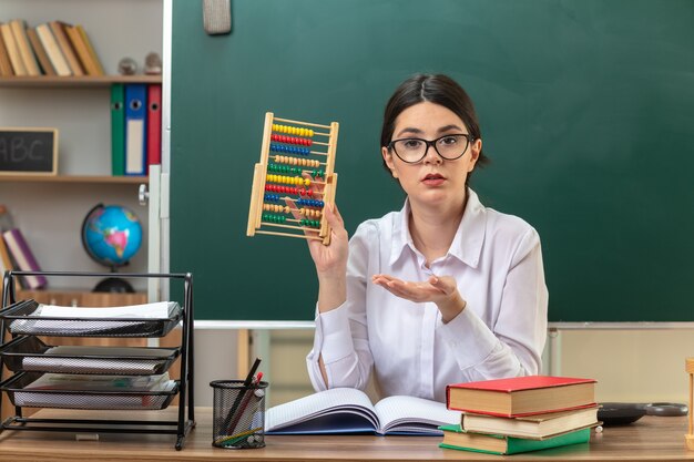 Confused young female teacher wearing glasses holding and points with hand at abacus sitting at table with school tools in classroom
