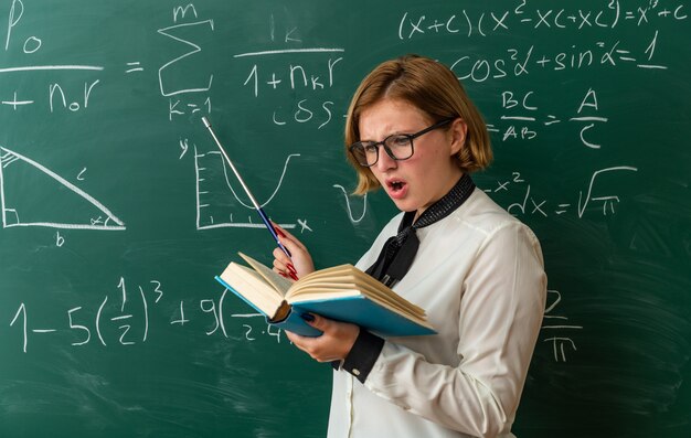 Confused young female teacher wearing glasses standing in front blackboard reading book in classroom