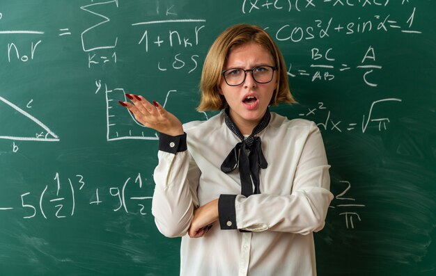 Confused young female teacher wearing glasses standing in front blackboard spreading hand in classroom