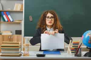 Free photo confused young female teacher wearing glasses tear paper sitting at desk with school tools in classroom