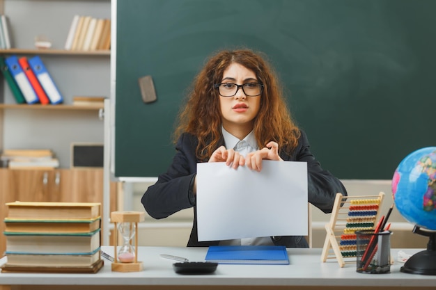 教室で学校の道具と机に座って眼鏡の涙の紙を身に着けている混乱した若い女性教師