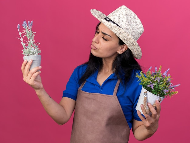 Confused young gardener girl wearing uniform and hat holding and looking at flowerpots isolated on pink wall