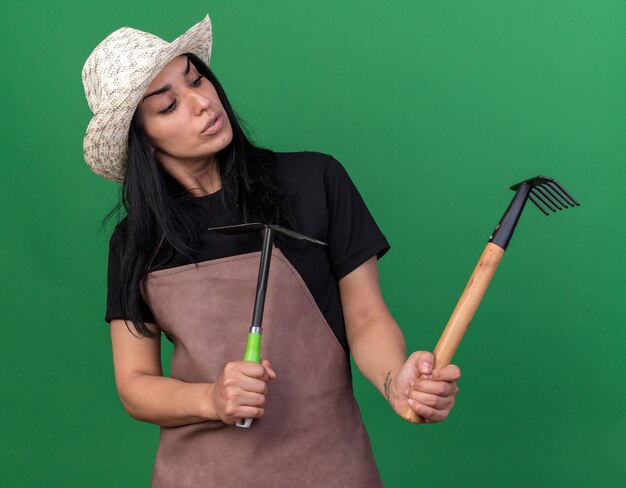 Confused young gardener girl wearing uniform and hat holding rake and hoe-rake looking at rake isolated on green wall