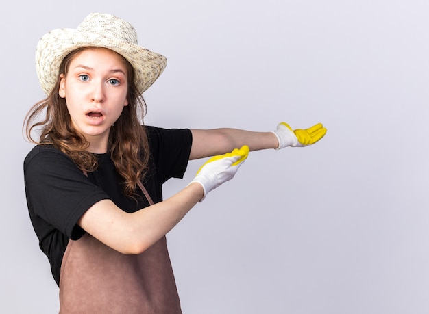 Confused young gardener wearing gardening hat with gloves points at behind 