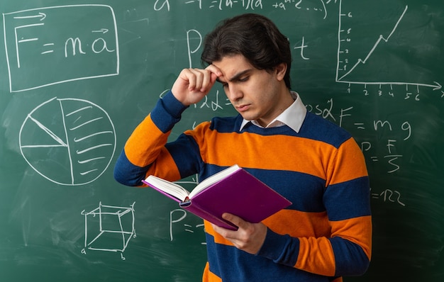 Foto gratuita confuso giovane insegnante di geometria in piedi davanti alla lavagna in aula toccando il libro di lettura sulla fronte