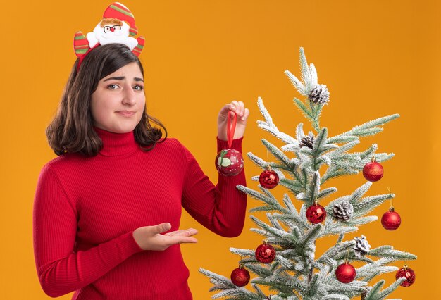 오렌지 배경 위에 크리스마스 트리 옆에 재미있는 머리띠를 입고 크리스마스 스웨터에 혼란 어린 소녀