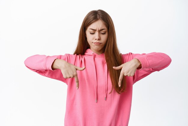 Confused young girl having doubts frowning and pointing down looking with hesitant unsure face standing troubled against white background