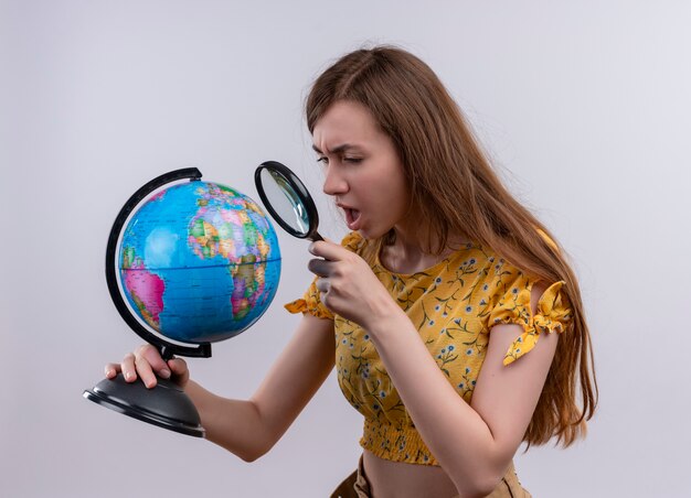 Confused young girl holding globe and looking through magnifier glass on isolated white space