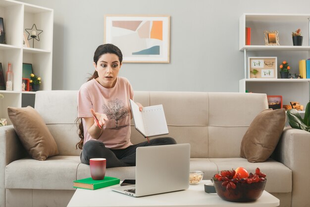 Confused young girl holding notebook sitting on sofa behind coffee table looking at laptop in living room