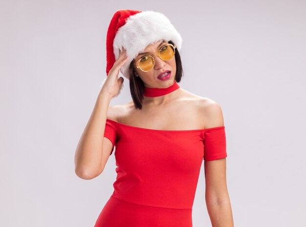 Confused young girl wearing santa hat and glasses looking at camera touching head isolated on white background