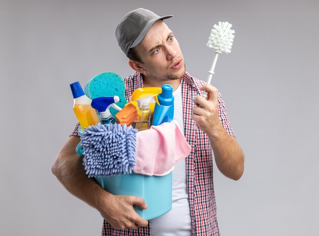 confused young guy cleaner wearing cap holding bucket with cleaning tools and looking at brush in his hand isolated on white wall