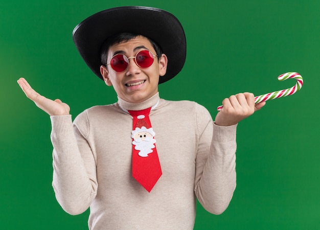 Confuso giovane ragazzo che indossa il cappello con cravatta natalizia e vetri che tengono le mani di diffusione della caramella di natale isolate sulla parete verde