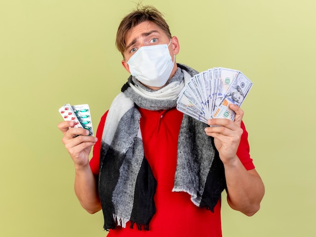 Confused young handsome blonde ill man wearing mask holding money and packs of medical pills looking at front isolated on olive green wall